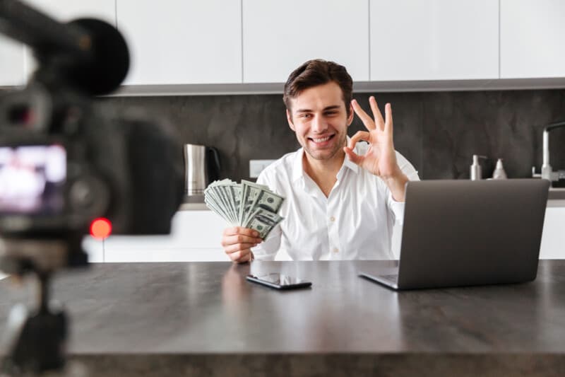 Cheerful young man filming his video blog episode about new tech devices while sitting at the kitchen table with laptop and showing bunch of money banknotes