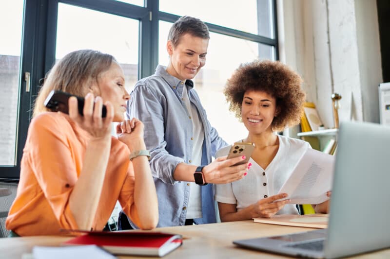 A group of diverse female professionals gathered around a table, engrossed in a cell phone.