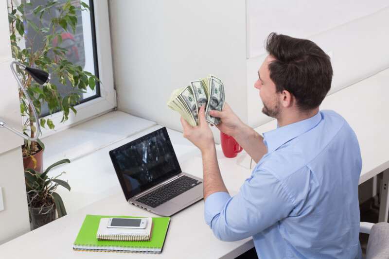Male freelancer counting money or dollars in office while working on laptop or computer. Handsome man looking on money